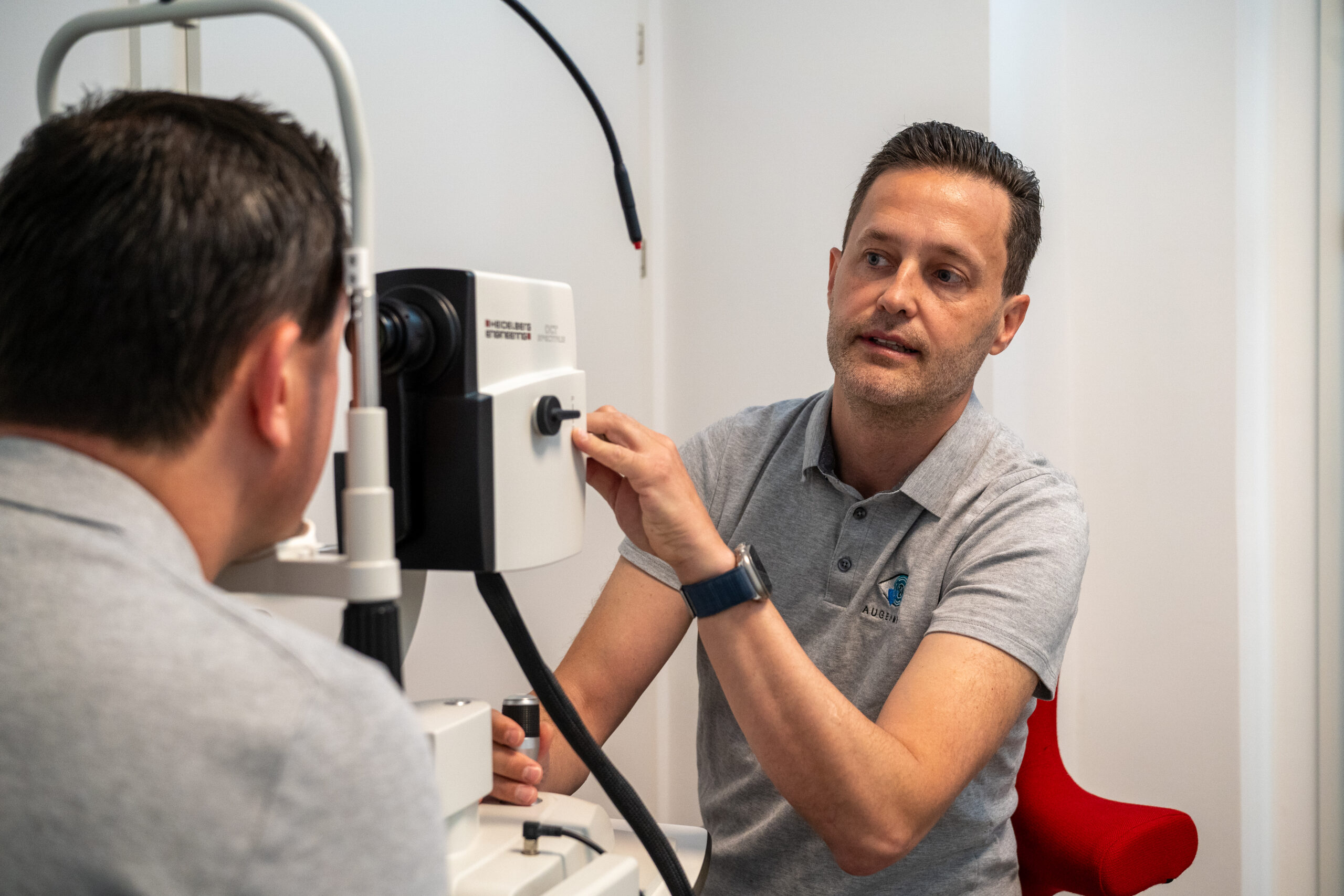 Prof. Dr. Mayer trägt ein graues Augen Mayer Poloshirt und sitzt vor einem Untersuchungsgerät. Er untersucht einen Patienten, um die Eignung für das Augenlasern zu überprüfen
