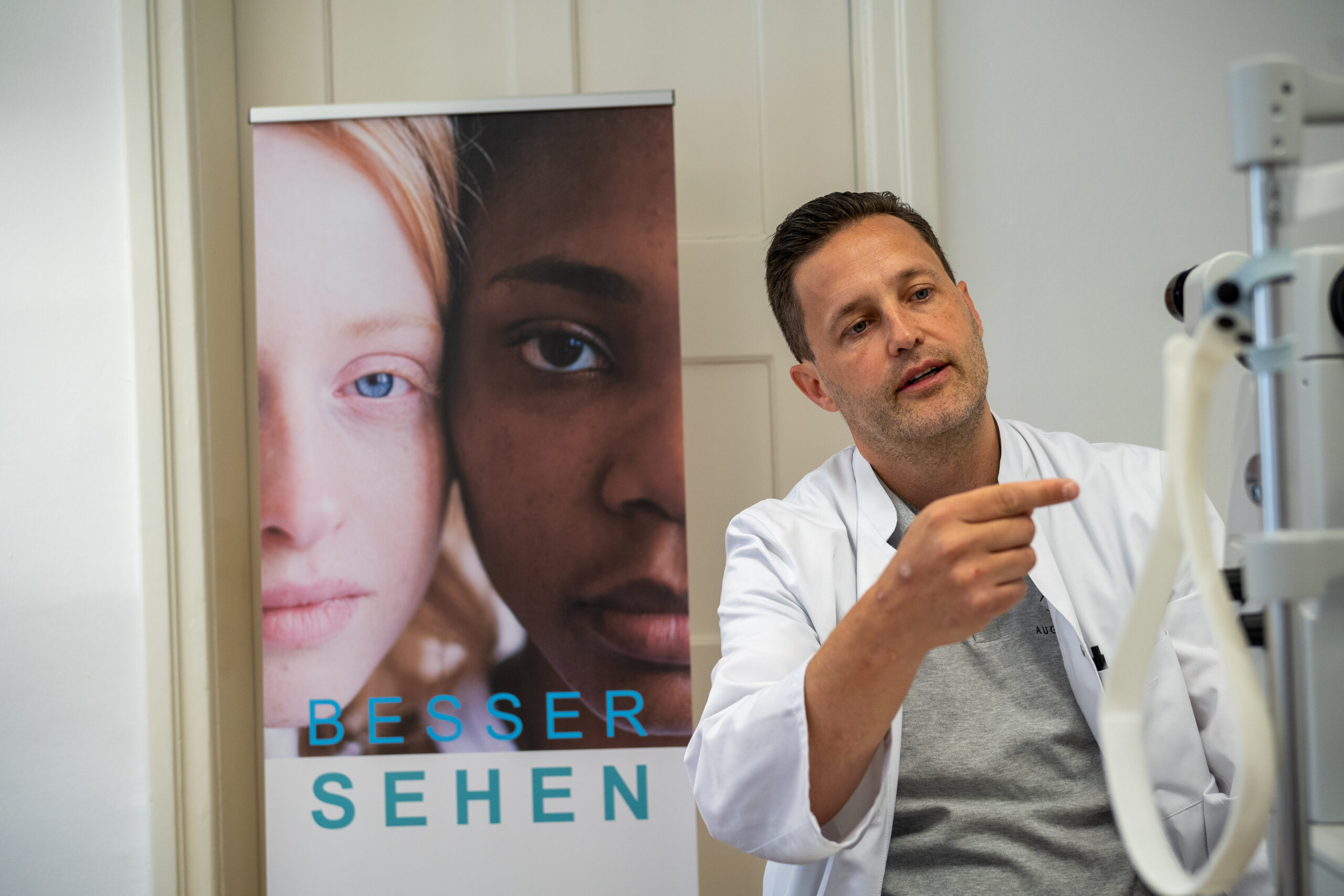 Prof. Dr. Mayer sitzt in einem Behandlungszimmer, trägt ein graues Polo und einen weißen Arztkittel. Er gestikuliert, während er einem Patienten die Vorteile einer Augenlaserbehandlung zeigt.